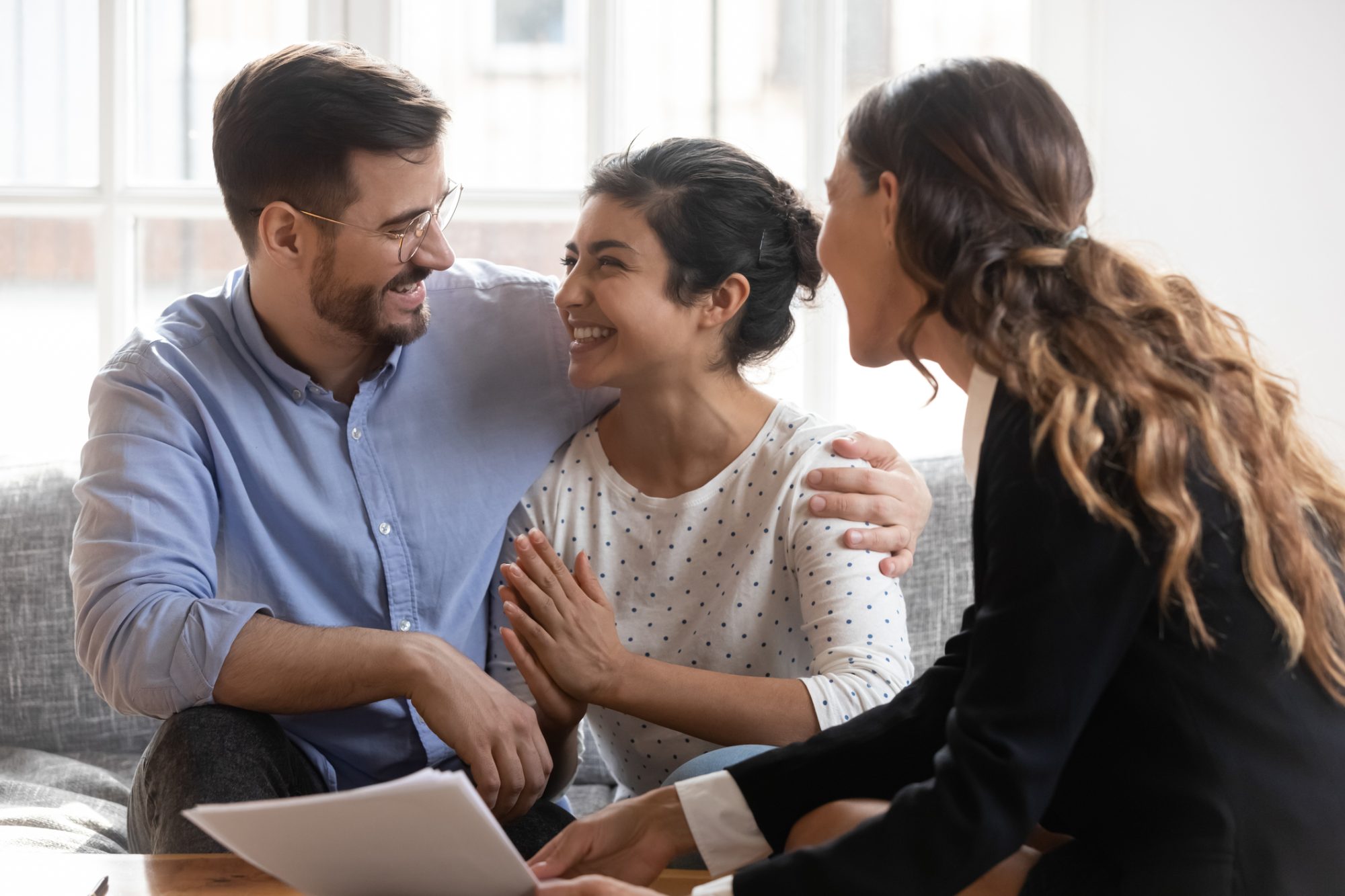 Happy diverse couple making decision at meeting with advisor, looking at each other, smiling Indian women and Caucasian men purchasing new apartment, taking loan or mortgage, satisfied clients