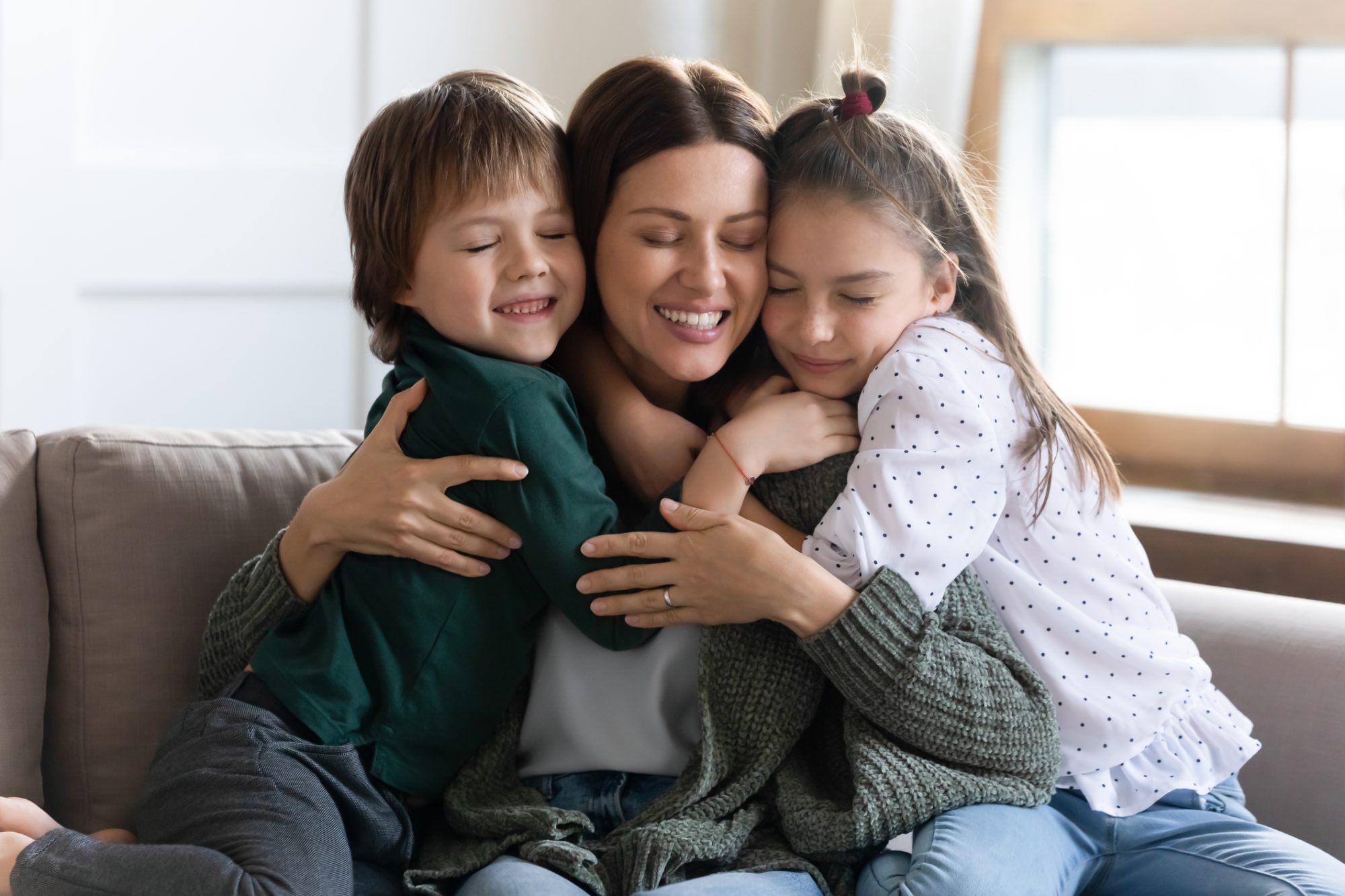 Affectionate small children embracing young mother, missing her after long separation. Loving mommy nanny babysitter cuddling little kids, relaxing together on comfortable couch in living room.