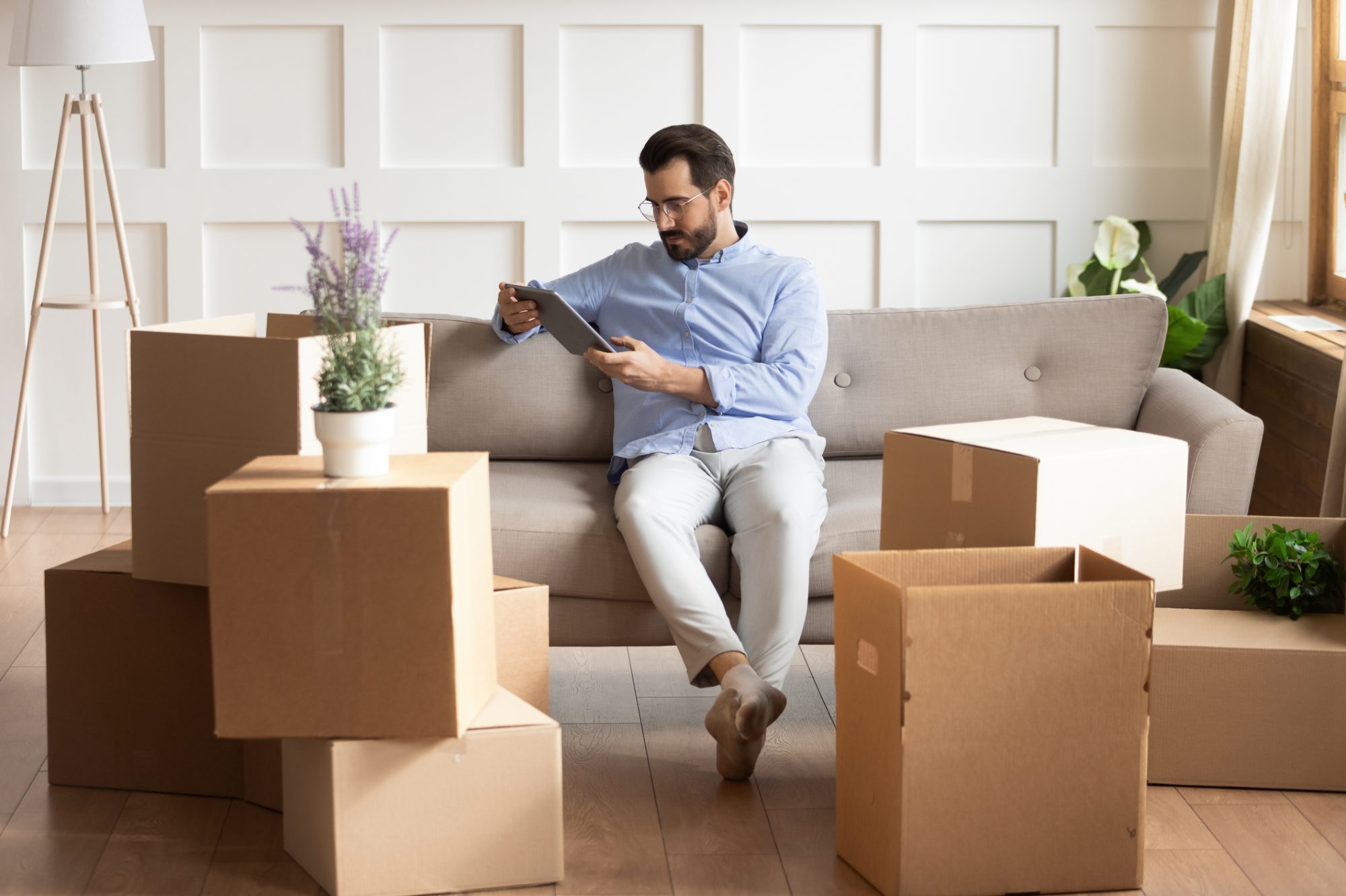 Young Caucasian man sit on couch relocate to new home relax using modern tablet gadget. Millennial male renter rest on moving day look at laptop screen browse surf wireless internet on pad device.